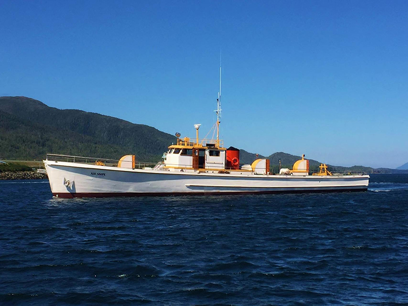 A long white boat named “Air Snipe” moves across open water with forested hills in the background under a clear sky.