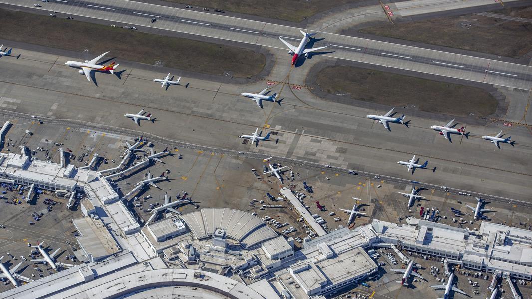 An aerial view of Sea Airport showing multiple commercial airplanes taxiing on parallel taxiways, with additional aircraft parked at terminal gates and airport buildings in the foreground.