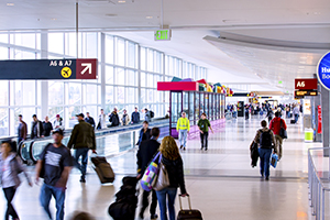 Interior of a busy airport concourse with travelers walking through the hallway, some carrying luggage. Overhead signs display gate information including A6 and A7.