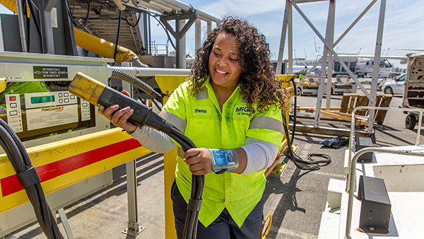 A person wearing a high‑visibility work shirt holds a large hose connector at an outdoor equipment station, with aircraft and service vehicles visible in the background.