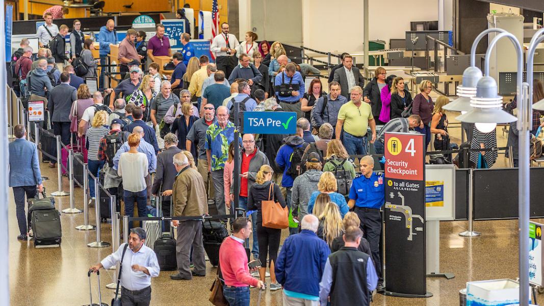 A large crowd waits in multiple lines at an airport security checkpoint, with signs for TSA Pre✓ and Checkpoint 4 visible.