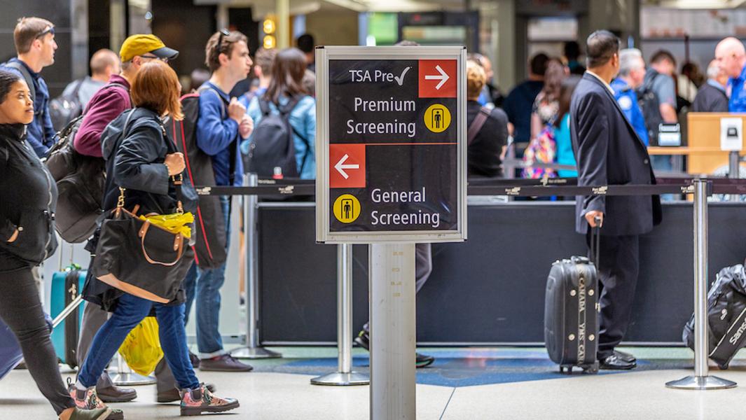 A sign in an airport directs travelers to Premium Screening on the right and General Screening on the left, with people walking and waiting in lines behind it.