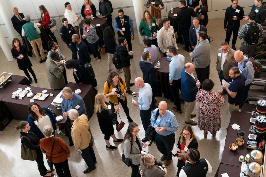 People gathered in a large indoor space, standing in small groups and talking during a networking event with tables and refreshments nearby.