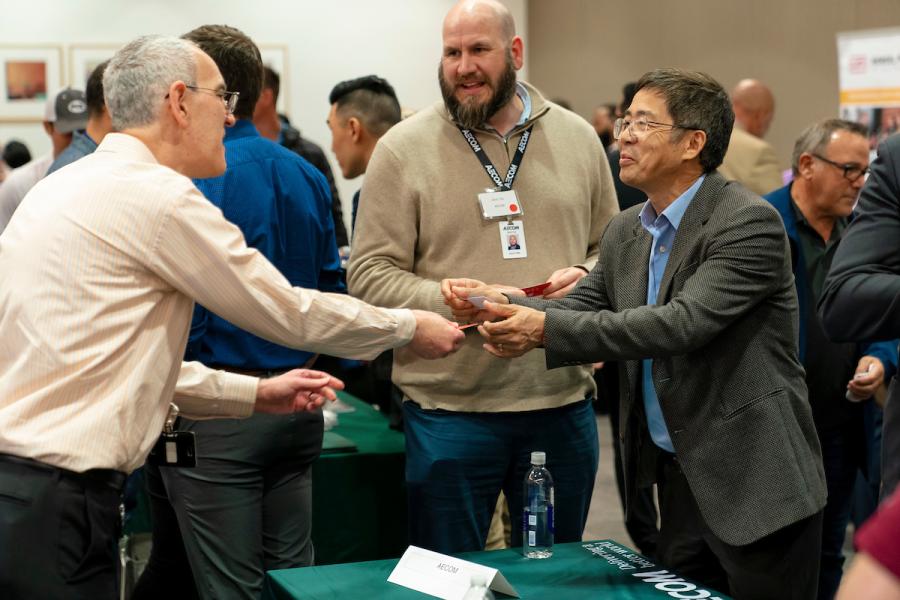People interacting at a table during a networking event, with two individuals exchanging items while others stand nearby.