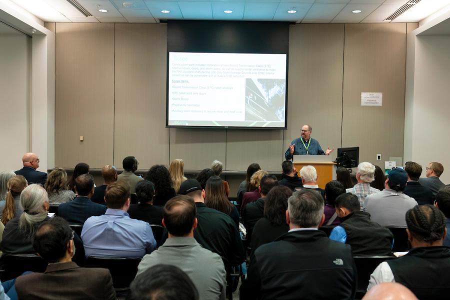 An audience seated in a conference room listening to a speaker presenting at a podium with a slide projected on a screen.
