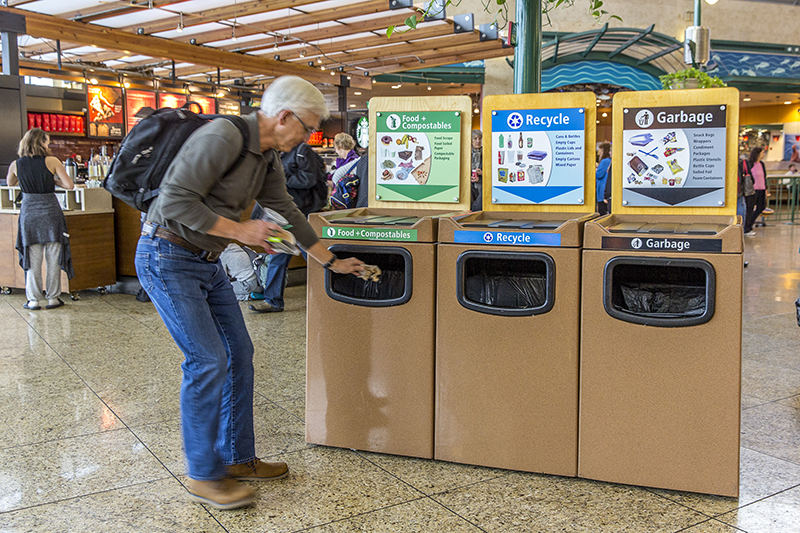 A person places an item into a food‑and‑compostables bin beside recycle and garbage bins in a public indoor area.