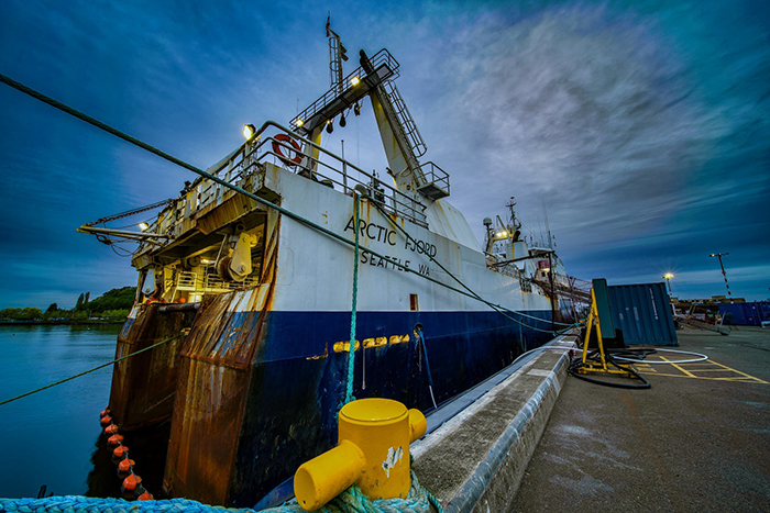 A large fishing vessel docked at a pier in low evening light, with ropes securing it and cloudy sky overhead.