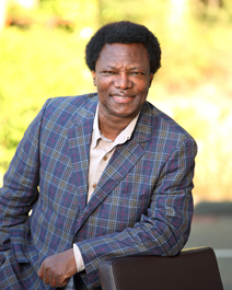 Ato Apiafi A person wearing a patterned jacket leans on a chair outdoors with greenery in the background.