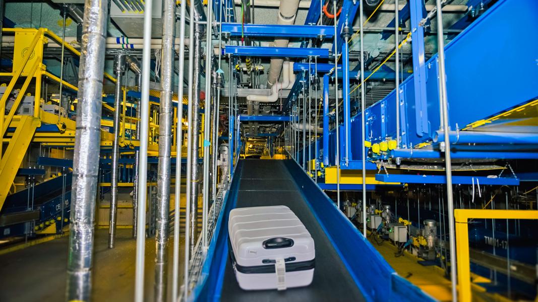 A suitcase moves along a conveyor belt inside SEA Airport’s automated baggage system, surrounded by blue and yellow machinery, pipes, and support structures.