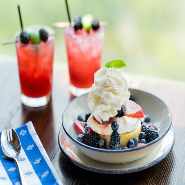 A bowl of shortcake topped with whipped cream and assorted berries sits on a wooden table, with two red berry drinks in the background.