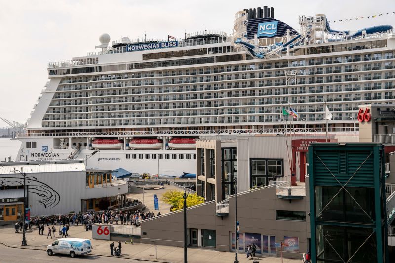 The cruise ship Norwegian Bliss is docked beside the Bell Harbor complex, with passengers lined up near the Pier 66 entrance.