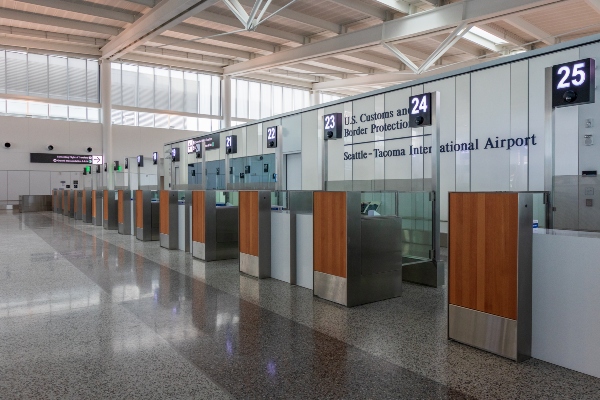 An empty U.S. Customs and Border Protection inspection area at Seattle–Tacoma International Airport shows a row of numbered processing booths with glass partitions and wood panels, lit by natural light from large windows above.