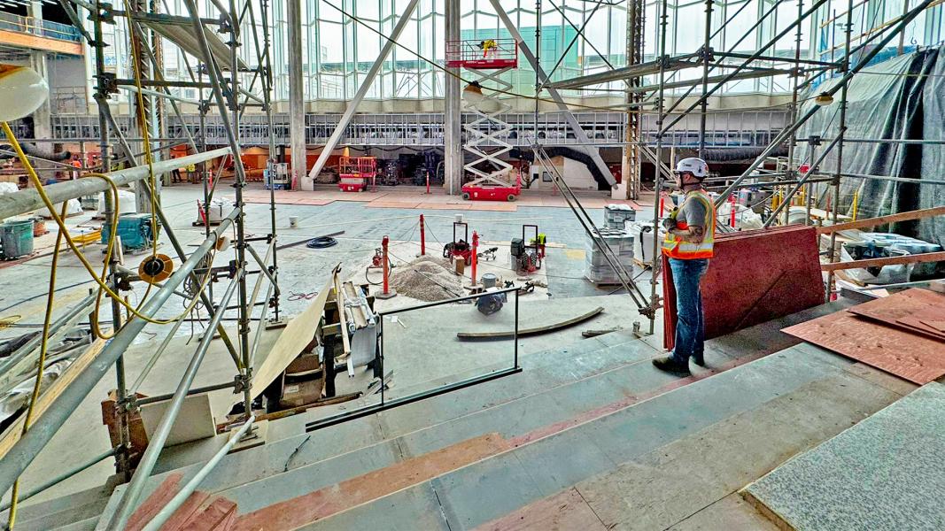 A worker in safety gear stands inside the C Concourse Expansion construction area at SEA Airport, surrounded by scaffolding, equipment, and building materials.
