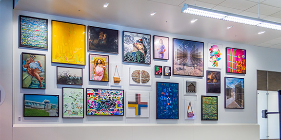 A large indoor wall displays an eclectic arrangement of framed artworks in various sizes, styles, and colors, including abstract designs, portraits, landscapes, geometric patterns, and mixed‑media pieces, all mounted above a white lower wall panel.