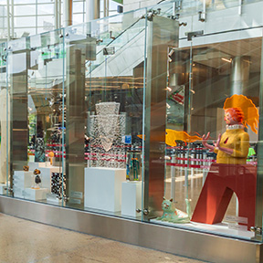 A glass display case in a bright indoor area showcases several sculptural pieces, including a tall abstract form made of layered materials and a large colorful cutout figure with exaggerated hair and extended arms. Additional small artworks sit on white pedestals behind the glass.