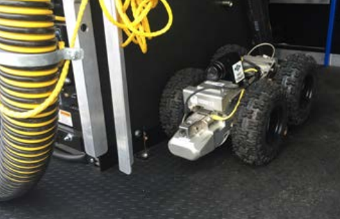 A wheeled inspection robot with a mounted camera and thick tires positioned beside cables and equipment inside a work vehicle.