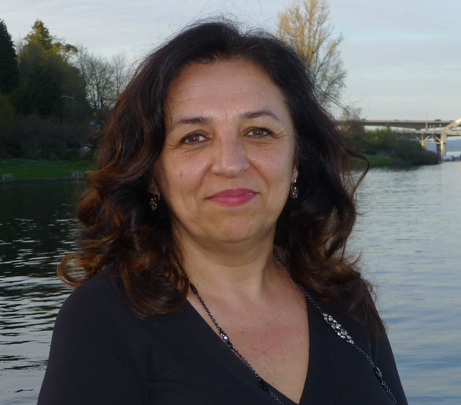 Bookda Gheisar standing outdoors near a body of water, with trees and a bridge in the background.