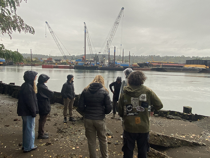 A small group of people stand on a rocky shoreline facing a river, with cranes, barges, and industrial structures visible across the water on an overcast day.