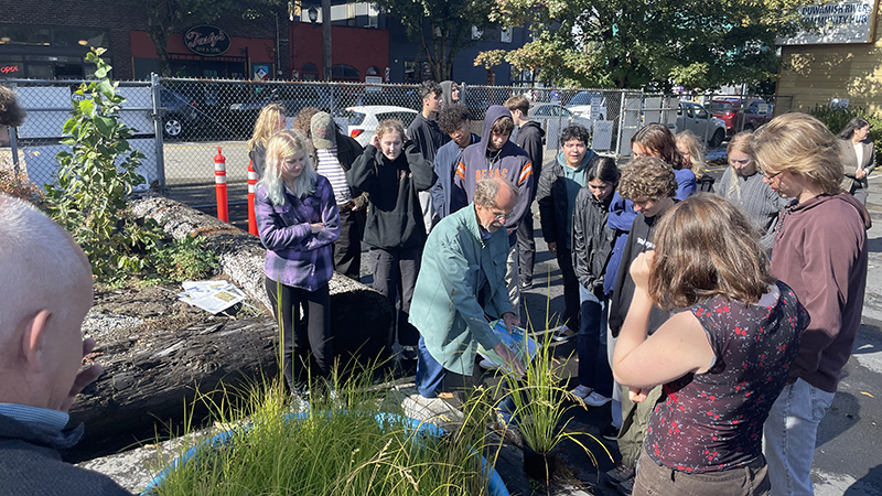 A group of people gather outdoors around a person demonstrating how to handle plants in a small garden area, with a fence, nearby buildings, and sunlight filtering through trees in the background.