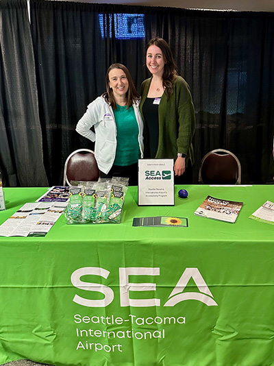 Two people standing behind a SEA Airport outreach table with brochures, small giveaway items, and a sign labeled “SEA Access.