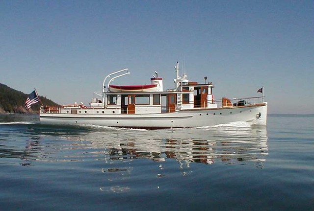 A white boat named “Carmelita” moves across calm water, with wood‑trimmed cabin structures and an American flag at the stern, and forested shoreline visible in the background under a clear sky.