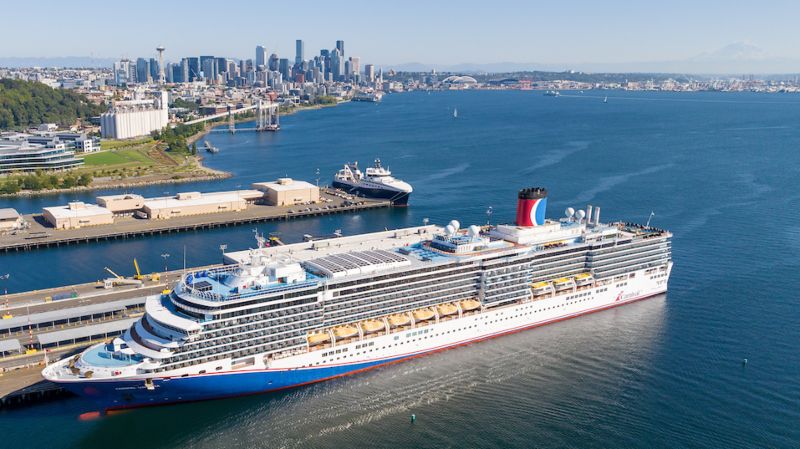 A cruise ship moves across a wide bay with downtown Seattle in the background, including the Space Needle on the left side of the skyline.