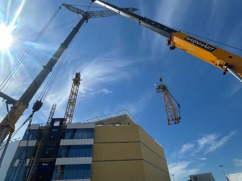 A mobile crane removes a section of a tower crane beside a building under a clear sky.