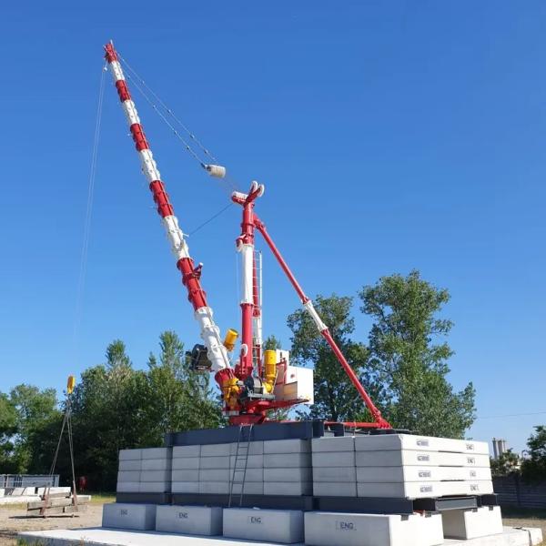 A red‑and‑white crane stands on stacked counterweight blocks outdoors under a clear blue sky.