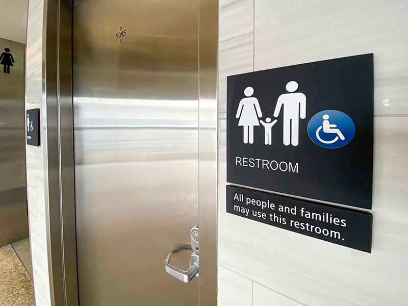A restroom entrance with a metal door and a sign showing restroom symbols, an accessibility symbol, and text stating that all people and families may use the restroom.