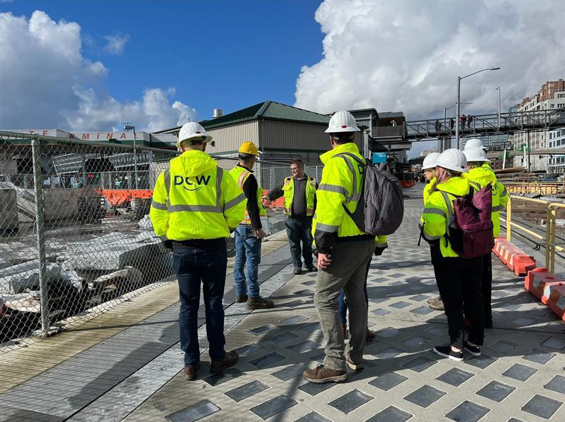 Workers wearing high‑visibility safety gear and hard hats gather on a construction site near fencing and industrial buildings.