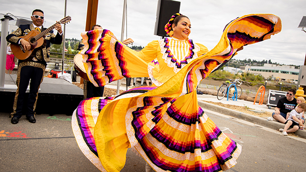 A dancer in a bright, multicolored traditional-style dress spins outdoors while a musician plays guitar nearby.