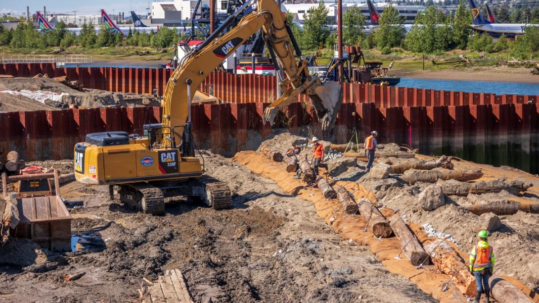 Construction equipment and workers place large logs and materials along a shoreline area inside a steel sheet‑pile enclosure, with water and industrial buildings in the background.