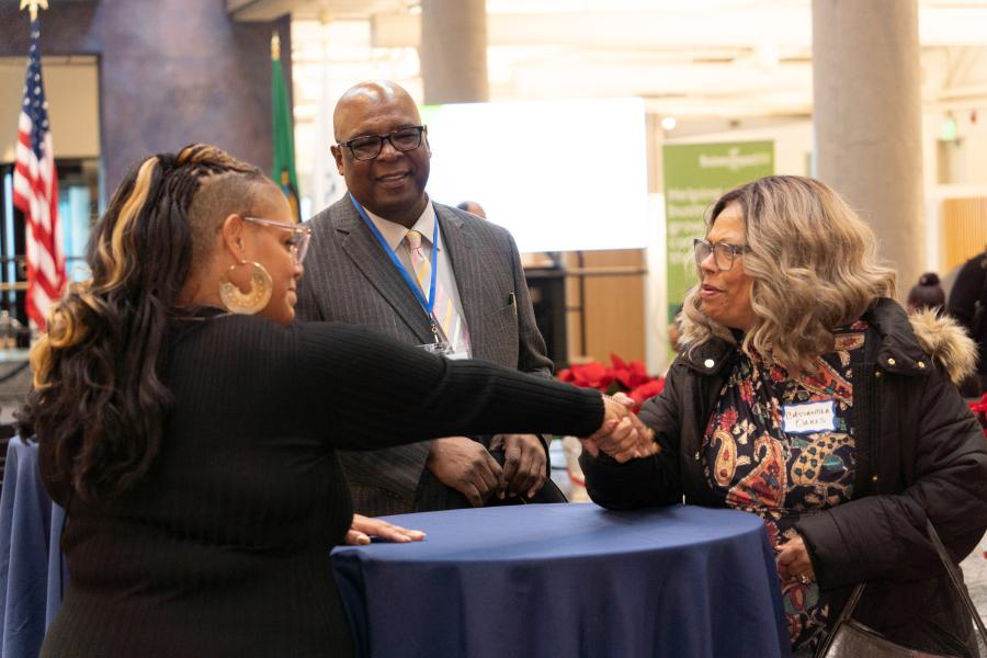 Two people shake hands at a round table while another person stands nearby in an indoor event space.