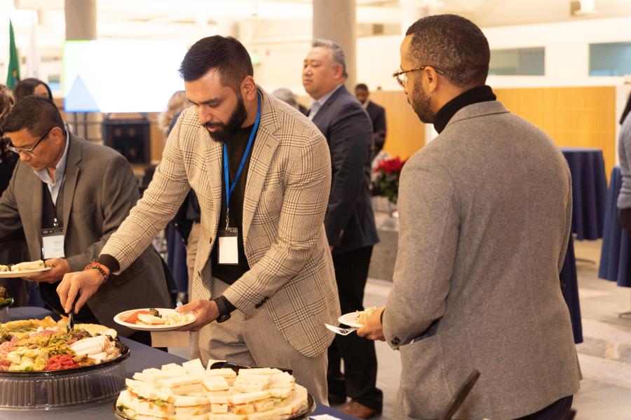 A group of people stand at a buffet table in an indoor event space. Several individuals are selecting food from trays filled with sandwiches, vegetables, and other appetizers. The setting includes tables covered with blue tablecloths and a background with additional attendees and event furnishings.