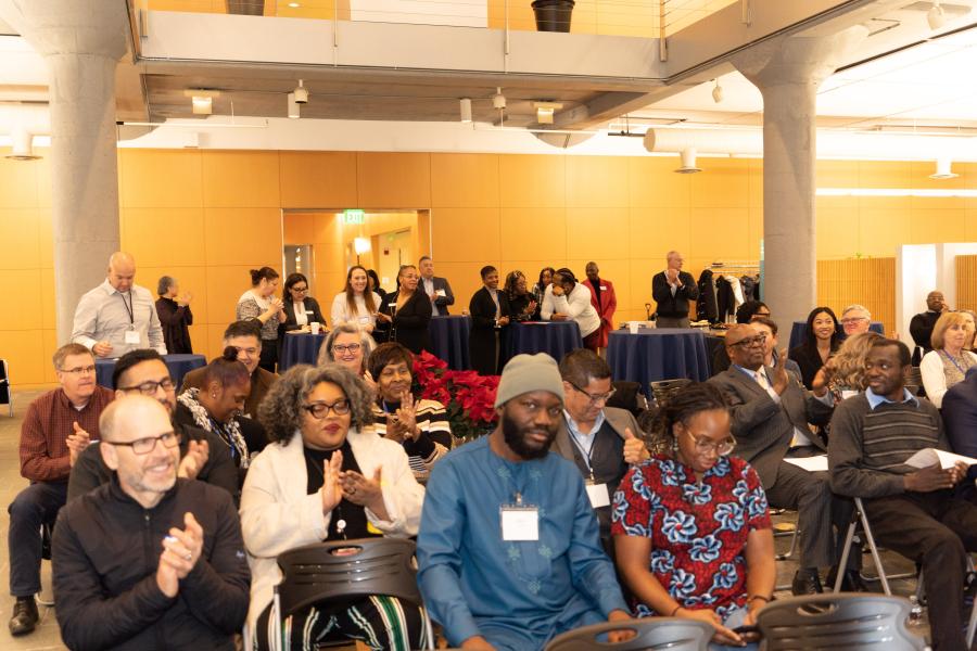 An indoor event audience sits in rows while additional attendees stand near tables in the back of the room.