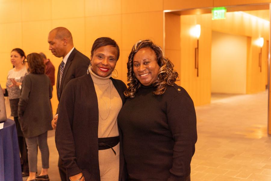 Darci and Tamaka Thornton standing together and posing in a warmly lit indoor space, with several others talking in the background.