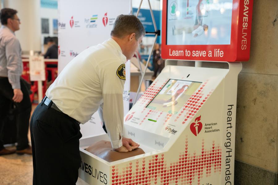 Chief Krause practices hands‑only CPR on an American Heart Association training kiosk in a public indoor area.