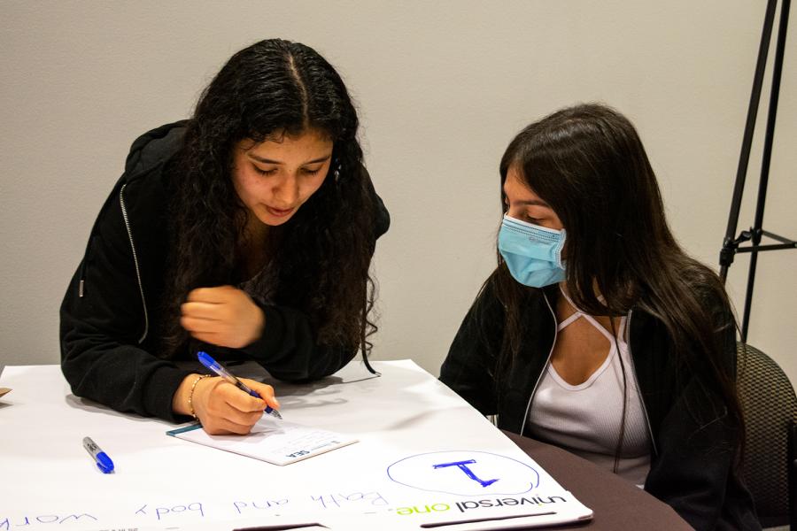 Two individuals sit at a table working on a large sheet of paper, with one individual writing and the other watching.