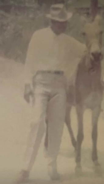 A vintage photo of a Daddy ST wearing a hat and light clothing walking beside a horse on a dirt path.