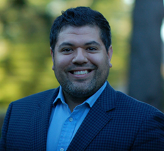 David Molina  wearing a checkered blazer and a collared shirt stands outdoors with blurred greenery in the background, looking toward the camera.
