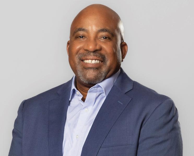 Delmas Whittaker wearing a suit jacket and collared shirt poses for a head‑and‑shoulders portrait against a plain background.