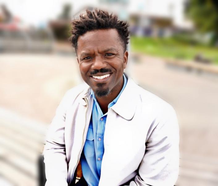Delton Johnson wearing a light jacket and a button‑up shirt sits outdoors with a blurred park-like background.
