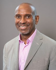 Dexter Turner wearing a light checkered jacket and a pink collared shirt stands against a plain gray background, looking toward the camera.