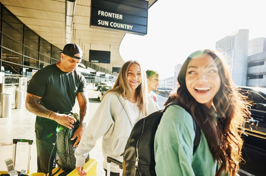People walking with luggage outside an airport terminal under bright sunlight. Overhead signs show airline names, and vehicles are parked along the curb.