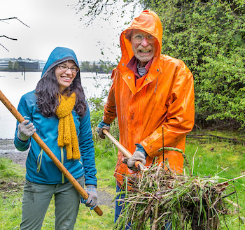 George Blomberg in high-visibility vest using a clipboard at a shoreline habitat restoration site near Seattle, with water and native plants visible