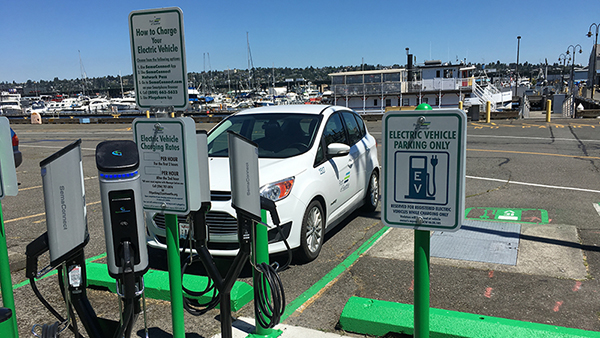 Electric vehicle charging stations stand in a parking area near a waterfront, with a white car parked in an EV‑designated space.