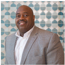 Edward Byers wearing a light-colored checkered jacket and a white collared shirt stands in front of a patterned teal-and-white wall, looking toward the camera.