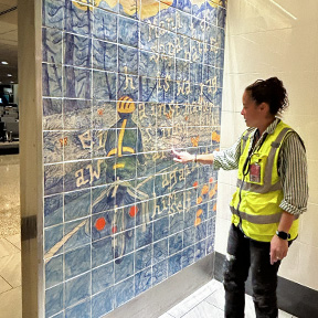A person wearing a safety vest stands in front of a tiled mural inside an indoor walkway, gesturing toward artwork that depicts a figure on a path with blue and yellow tones and scattered text across the tiles.