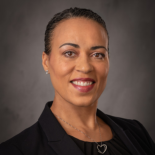 Erica Norton wearing a dark blazer and a necklace with a heart-shaped pendant poses against a dark, softly textured background.