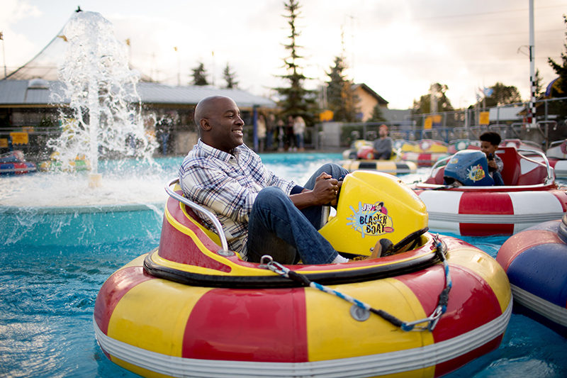 A person sits in a circular bumper boat on a pool, holding the steering controls, with water spraying from a fountain in the background and other bumper boats and onlookers visible around the attraction.